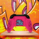 "Custom Arcane inspired Xbox Series X controller with vibrant graffiti art design, featuring bright blue, pink, and purple street-style graphics with 'BOOM' text on the touchpad, displayed in front of an urban graffiti wall."