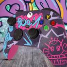 "Custom Arcane inspired Xbox Series X controller with vibrant graffiti art design, featuring bright blue, pink, and purple street-style graphics with 'BOOM' text on the touchpad, displayed in front of an urban graffiti wall."